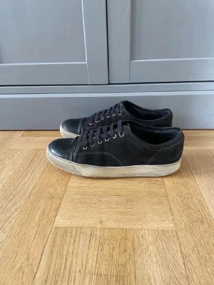 Svart Lanvin Cap-Toe (Stl 43/UK9) - Svarta glansiga Lanvin cap-toe sneakers, storlek UK 9 (EU 43). Skick: bra – lätt slitage fram på tån som syns på bilderna, i övrigt fräscha. Säljs utan box. Hör av dig vid frågor!