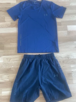 Blått träningsset från Under Armour - Snyggt blått träningsset från Under Armour med t-shirt och shorts. T-shirten har diskret logga på bröstet och text längs sidan. Shortsen har elastisk midja, logga på benet och ventilerande material. Perfekt för gym eller löpning.