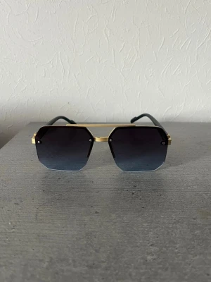 Solglasögon  - Snygga solglasögon med kantig form och svarta glas som tonas mot blått. Bågarna har guldfärgade detaljer och en modern, stilren look. Perfekta för dig som vill sticka ut med accessoarer som känns lyxiga och trendiga.