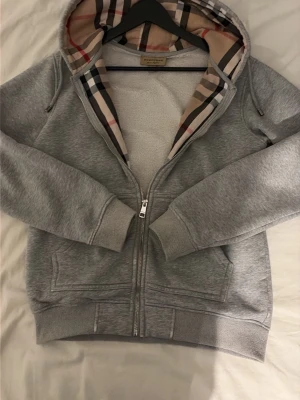 Grå zip hoodie från Burberry - Snygg grå zip hoodie från Burberry med klassiskt rutigt foder i huvan och diskret broderad logga på bröstet. Hoodien har dragkedja framtill, två fickor och ribbade muddar. Perfekt för en chill och stilren look. Pris kan diskuteras!