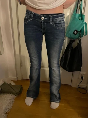 Blå bootcut jeans med kontrastsömmar - Säljer ett par blå bootcut jeans med vita kontrastsömmar och snygga detaljerade bakfickor med knappar. Jeansen har låg midja och klassisk femficksdesign. Perfekta för dig som gillar en avslappnad men ändå trendig stil❣️