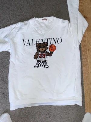 Vit Valntino sweatshirt med björn - Vit sweatshirt från Valntino med ett stort tryck av en björn i basketlinne och sneakers som håller en basketboll. Texten 'VALNTINO' står ovanför motivet. Tröjan har rund halsringning och långa ärmar. Perfekt för dig som gillar streetwear och coola prints.