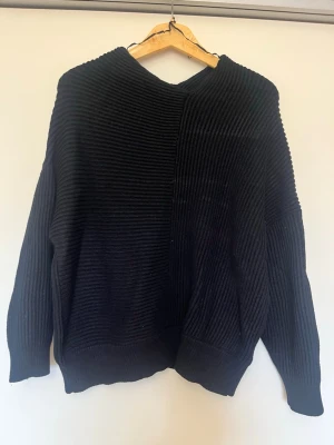 Svart ribbad stickad tröja - En svart stickad tröja med bred ribbad struktur och avslappnad passform. Tröjan har långa ärmar och en något boxig siluett, perfekt för lager på lager. Enkel och stilren design utan synliga loggor eller detaljer. oanvänd har bara hängt i garderoben 
