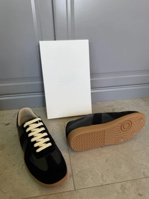 Svarta Maison Margiela - Stilrena svarta sneakers från Maison Margiela med kontrasterande vita skosnören och beige yttersula. Ovandelen är i en mix av mocka och läder för en lyxig känsla. Klassisk låg modell som passar till många olika outfits.