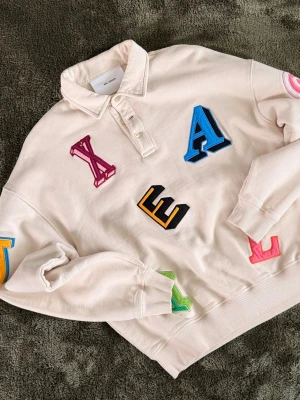Axel Arigato - Säljer en beige polotopp från Axel Arigato med färgglada bokstäver i olika stilar broderade över hela tröjan. Toppen har krage, knappar vid halsen och ribbade muddar vid ärmslut och nederkant. Perfekt för dig som gillar statement-plagg!
