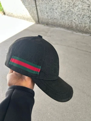 Svart keps från Gucci med logga - Säljer en svart keps från Gucci med diskret GG-mönster över hela kepsen och klassiska gröna och röda ränder på sidan. Kepsen har böjd skärm och justerbar rem bak. Materialet är bomull och insidan har Gucci-logga på banden.