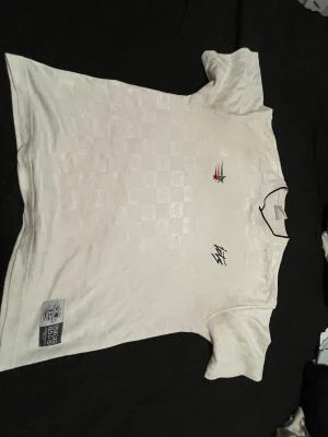 Vit retro fotbollströja med tryck - Säljer en vit retro fotbollströja från Le Coq Sportif med schackrutigt mönster i tyget, broderad logga och färgglada detaljer på bröstet. Nummer 17 i rött på ryggen och svart kant runt kragen. Perfekt för dig som gillar klassisk sportstil.