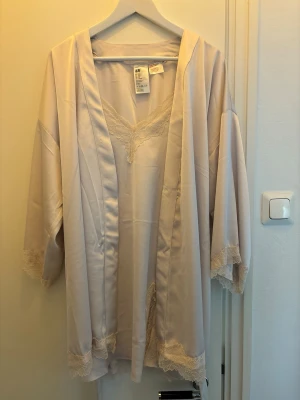 Ljusbeige morgonrock och nattlinne i satin med spets  - Elegant ljusbeige morgonrock/nattlinne från H&M i satinliknande material med fina spetsdetaljer vid ärmslut, fåll och halsringning. Morgonrocken har vida ärmar och ett matchande band i midjan för en avslappnad och lyxig känsla.