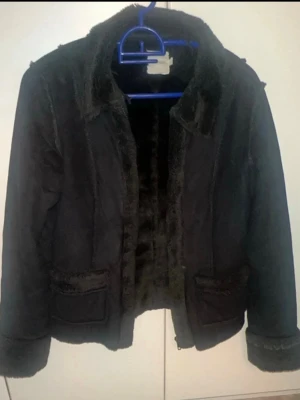 Svart pälsjacka med fuskpälsdetaljer - Snygg svart pälsjacka med mjuk fuskpäls på insidan, krage och ärmslut. Jackan har två fickor framtill och markerade sömmar för en cool look. Perfekt för kalla dagar när du vill vara både varm och trendig.