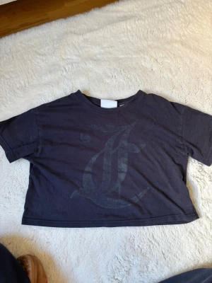 Svart croppad t-shirt Juicy Couture - Snygg svart croppad t-shirt från Juicy Couture med ett stort ton-i-ton tryck framtill. Klassisk rund halsringning och korta ärmar. Perfekt för en avslappnad och trendig look.
