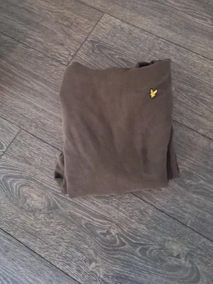 Olivgrön hoodie från Lyle & Scott - Säljer en olivgrön hoodie från Lyle & Scott med klassisk gul logga på bröstet. Hoodien har huva och en stor magficka framtill. Tillverkad i mjukt bomullsmaterial och har en avslappnad passform. Perfekt för dig som gillar stilrena och bekväma plagg.