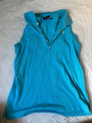 Turkos ärmlös pikétopp från Ralph Lauren - Snygg turkos ärmlös pikétopp från Ralph Lauren med klassisk krage och knappar framtill. Toppens material är bomull och den har det ikoniska gula broderade logotypen på bröstet. Perfekt för en fräsch och sportig look.