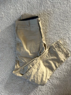 Beige chinos från Bläck, tailored fit - Snygga beige chinos från Bläck i modellen tailored fit. Byxorna har klassisk design med fickor fram och bak, samt bälteshällor. Perfekta för en clean och stilren look. Tillverkade i mjukt och bekvämt material som passar året runt.
