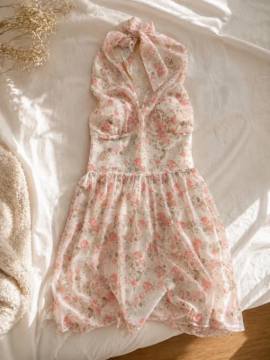 Blommig halterneckklänning i chiffong - Söt kortklänning med halterneck och knyt i nacken. Klänningen är vit med rosa och gröna blommor, har v-ringning och volangig kjol. Tillverkad i ett tunt, lätt material som ger en romantisk känsla.