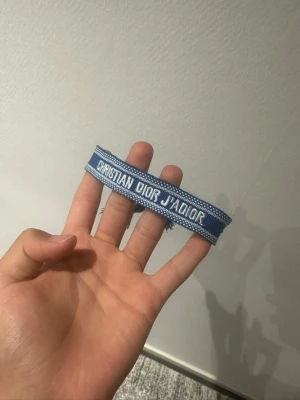 Blått tygarmband Christian Dior - Snyggt blått tygarmband från Christian Dior med texten 'CHRISTIAN DIOR J'ADIOR' i vitt. Armbandet har justerbar knytning med tofsar i ändarna och en stilren, ungdomlig design. Perfekt accessoar för att lyfta din outfit.