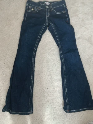 Mörkblå bootcut jeans Gina Tricot - Snygga mörkblå bootcut jeans från Gina Tricot med kontrastsömmar och klassisk femficksmodell. Jeansen har vita och röda detaljer vid sömmarna och en knapp med logga framtill. 
