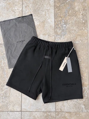 Svarta Essentials shorts med snörning - Svarta shorts från Essentials med diskret logga på vänster ben. De har elastisk midja med snörning och en relaxed passform. Tillverkade i mjukt bomullsmaterial, perfekta för chill dagar eller streetwear-stil. Kommer med originalpåse och taggar kvar.