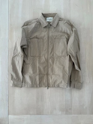 Beige overshirt - Stilren beige overshirt från  i tunn manchester med diskreta ränder. Två stora bröstfickor, dragkedja framtill och klassisk krage. Perfekt lager på lager-plagg med avslappnad passform och långa ärmar.