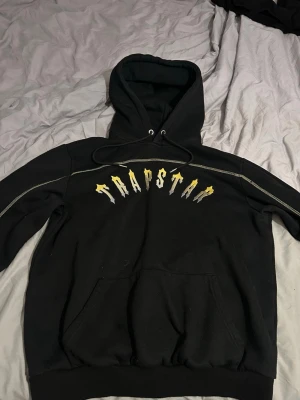 Svart Trapstar hoodie med tryck - Säljer en svart hoodie från Trapstar med stort gult och vitt tryck över bröstet. Hoodien har huva med dragsko, magficka och kontrastsömmar längs ärmarna. Perfekt för dig som gillar streetwear och vill ha en snygg och bekväm hoodie.