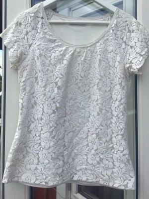 Vit blommig spetsblus - Söt vit blus med blommigt spetsmönster och korta ärmar. Toppen har rund halsringning och är tillverkad i ett genombrutet spetsmaterial som ger en romantisk vibe. Perfekt att styla med jeans eller kjol för en fräsch look.