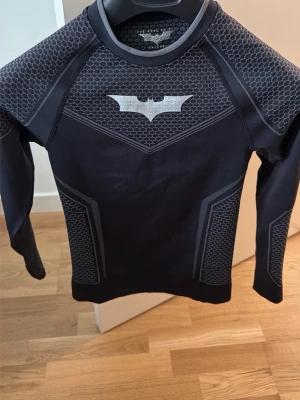Svart Batman långärmad topp - Svart långärmad topp från youngla med grå Batman-logga på bröstet och coola mönstrade detaljer på ärmar och sidor. Tillverkad i stretchigt syntetmaterial som sitter tight och ger en sportig vibe.