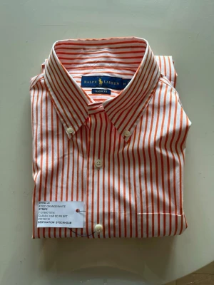 Randig skjorta från Ralph Lauren - Snygg skjorta från Ralph Lauren i klassisk passform med orange och vita vertikala ränder. Skjortan har button-down krage, långa ärmar och vita knappar framtill. Tillverkad i mjuk bomull som känns skön mot huden. Perfekt för en stilren och fräsch look.