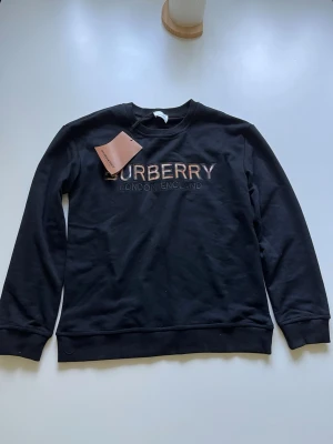 Svart sweatshirt från Burberry - Svart sweatshirt från Burberry med broderad logga 'BURBERRY LONDON ENGLAND' på bröstet. Tröjan har rund halsringning, långa ärmar och ribbade muddar. Tillverkad i mjukt bomullsmaterial för en bekväm känsla.