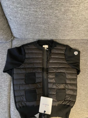 Svart dunväst från Moncler - Snygg svart dunväst från Moncler med quiltad framsida, fyra fickor och ribbade detaljer vid hals och ärmslut. Västen har dragkedja framtill och Moncler-logga på ärmen. Perfekt för lager-på-lager och streetstyle.! NFC finns.! Storleken är 1 vilket motsvarar S