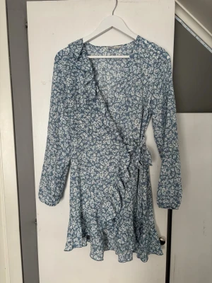 Blommig omlottklänning med volang - Använd en gång, som ny. Jätte skönt material då de är delvis stretch. Superfin omlottklänning med vitt blommönster och volangdetaljer längs kanten.  Klänningen har långa ärmar och knytband i midjan för en snygg passform. Perfekt för vår och sommar.  