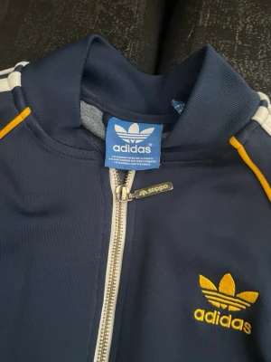  Adidas track jacket - Snygg marinblå track jacket från Adidas med dragkedja, vita ränder på axlarna och gula detaljer. Klassisk Adidas-logga broderad i gult på bröstet. Tillverkad i ett mjukt och stretchigt material som passar perfekt till träning eller streetwear.