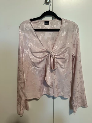 Ljusrosa blus med knytning från Gina Tricot - Säljer en lätt ljusrosa blus från Gina Tricot med vacker glansig finish och subtilt blommönster. Blusen har lång ärm och en snygg knytning framtill som ger en cool look. Materialet känns mjukt och lyxigt, perfekt för att lyfta din outfit.