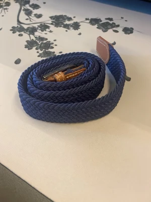 Mörkblå flätat bälte med läderdetaljer - Snyggt mörkblått flätat bälte med bruna läderdetaljer vid spännet. Bältet har en klassisk design och är gjort i ett flexibelt syntetmaterial som ger en bekväm passform. Perfekt accessoar för att lyfta din outfit.