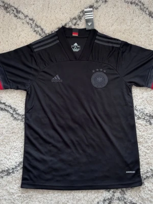 Svart Tyskland fotbollströja Adidas - Snygg svart fotbollströja från Adidas med Tysklands landslagslogga och fyra stjärnor på bröstet. Tröjan har korta ärmar, diskreta grå ränder på axlarna och röda detaljer vid ärmsluten. Tillverkad i lätt och ventilerande material med Aeroready-teknologi.