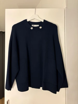 Mörkblå cape-kappa från Zara - Stilren mörkblå cape-kappa från Zara med två stora fickor framtill och dekorativa silverfärgade knappar vid halsen. Kappan har en rak, lös passform och är tillverkad i ett mjukt, tjockt tyg som ger en elegant look.