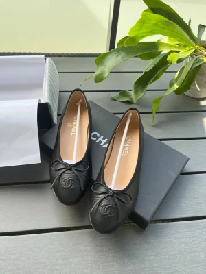 Svarta ballerinaskor från Chanel - Stilrena svarta ballerinaskor från Chanel i mjukt läder. Skorna har rund tå, dekorativ rosett framtill och den klassiska Chanel-loggan präglad på tån. Perfekta för dig som gillar tidlös och lyxig design.