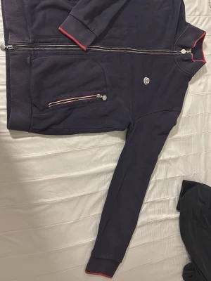 Mörkblå zip hoodie från Moncler - Snygg mörkblå hoodie från Moncler med dragkedja framtill och röda detaljer vid ärmslut och huva. Har fickor med dragkedja och Moncler-logga på bröstet. Tillverkad i mjuk bomull för skön känsla. Perfekt för en avslappnad och trendig stil. Finns ett hål som kanppt syns och den är exta finns bevis och en kurkod man skannar man går in i moncler om ni vill ha bild på de lila hålet kontakta mig säljer jättebilligt den är storlek L men kan användas som m ochså 