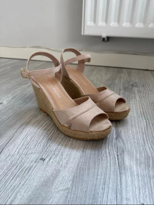 Beige kilklackade pumps Atmosphere - Snygga beige pumps från Atmosphere med öppen tå och kilklack i flätad jute. Skorna har en justerbar rem runt ankeln med guldfärgat spänne och ovandel i mockaimitation. 
