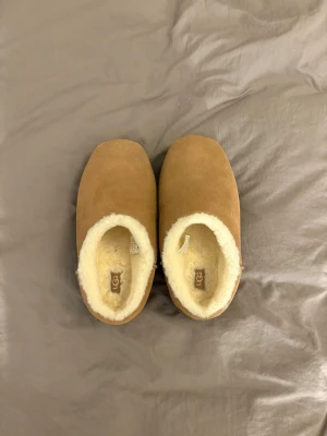 Bruna fårskinnstofflor från UGG - Mysiga bruna tofflor från UGG i äkta fårskinn med fluffigt vitt foder. Klassisk slip-in modell med rundad tå och mjuk innersula som håller fötterna varma. Perfekta för hemmabruk när du vill ha det extra bekvämt.