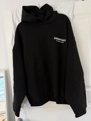 Svart Essentials Fear of God hoodie - Säljer en svart hoodie från Essentials Fear of God med vit logga på bröstet och stor text på ryggen. Hoodien har en rymlig huva och avslappnad passform. Perfekt för dig som gillar streetwear och vill ha en stilren look. Passar även dem som har storlek medium. Den är aldrig använd, endast upp packad.