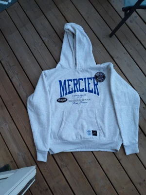 Grå hoodie från Mercier med tryck - Snygg grå hoodie från Mercier med blått tryck på bröstet och flera logodetaljer. Klassisk känguruficka framtill och ribbade muddar. Stor huva och mjukt material, perfekt för en avslappnad streetwear-look.