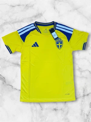 Sverige matchtröja från Adidas - Gul kortärmad matchtröja från Adidas med blå detaljer och Sveriges landslagsemblem på bröstet. Tröjan har blå ränder på axlarna och blå muddar vid ärmsluten. Tillverkad i ett lätt och ventilerande funktionsmaterial.