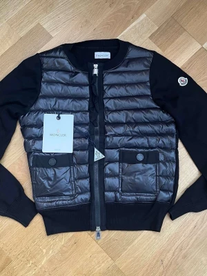 Svart  dam-cardigan från Moncler - Snygg svart cardiganjacka från Moncler med quiltad, glansig front och två fickor med knapp. Ärmar och rygg i mjuk stickad bomull, rund halsringning och dragkedja framtill. Ikonisk Moncler-logga på ärmen för extra stilpoäng. Kvitto finns även!!