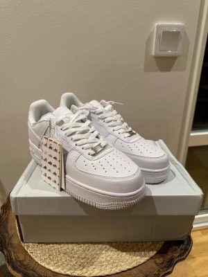 Nike Air Force 1 vita sneakers - Klassiska helvita Nike Air Force 1 sneakers i lågt utförande. Skorna har en stilren design med perforerad tå, vita skosnören och den ikoniska swoosh-loggan på sidan. Ovandelen är i slätt läder och sulan är tjock och greppvänlig. Perfekta för en clean och tidlös look.