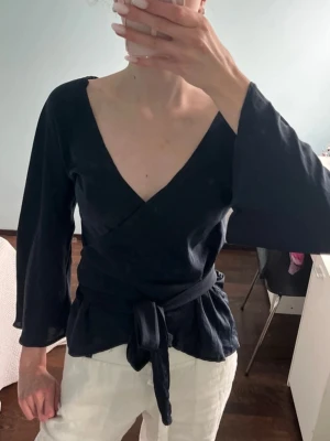 Navy omlottopp med knytband - Snygg svart omlottopp med djup v-ringning och knytband i midjan. Toppen har långa, lätt utsvängda ärmar och en feminin siluett. Perfekt att styla med både jeans och kjol för en trendig look.
