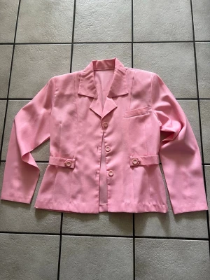 Rosa kavaj med knappar storlek M/L - Stilren rosa kavaj med klassisk krage och fyra ljusa knappar framtill. Kavajen har dekorativa knappar vid midjan och en bröstficka. Rak passform och långa ärmar. Perfekt för dig som vill sticka ut med färg.