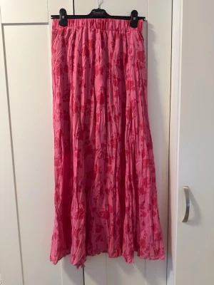 Rosa blommig långkjol - Säljer en rosa långkjol med blommigt mönster och plisserad design. Kjolen har elastisk midja och är luftig med ett flowigt fall, perfekt för dig som gillar färg och mönster. Materialet känns lätt och bekvämt.
