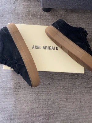 Svarta Axel Arigato sneakers i mocka - Snygga svarta sneakers från Axel Arigato i mjuk mocka med klassisk låg siluett och bruna gummisulor. Diskret logga på sidan och stilren design som passar till det mesta. Perfekta för dig som gillar minimalistisk och modern stil.