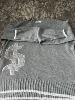 Grå stickad tröja från Many Studio - Snygg grå stickad tröja från Many Studio med vit dollar-tecken detalj på framsidan. Tröjan har ribbstickade muddar och nederkant med vita ränder. Perfekt för dig som gillar streetwear och vill sticka ut lite extra.