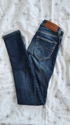 Jeans från Tiger of Sweden - Snygga blå jeans från Tiger of Sweden i klassisk femficksmodell. Jeansen har raka ben, kopparfärgade nitar och patch i brunt läder bak. Tillverkade i slitstark denim med tydliga sömmar och en tidlös tvätt. Säljer dessa då dem blivit alldeles för små för mig, hann bara användas en gång! Nypris 1700:-