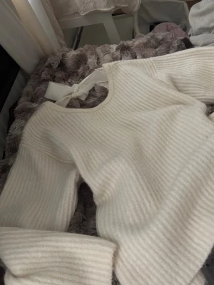 Vit ribbstickad tröja med rosett - Säljer en vit ribbstickad tröja med rund hals och en snygg rosett i nacken. Tröjan har långa ärmar och är gjord i ett mjukt, fluffigt material som känns riktigt cozy. Perfekt för dig som gillar stilrena och trendiga plagg.